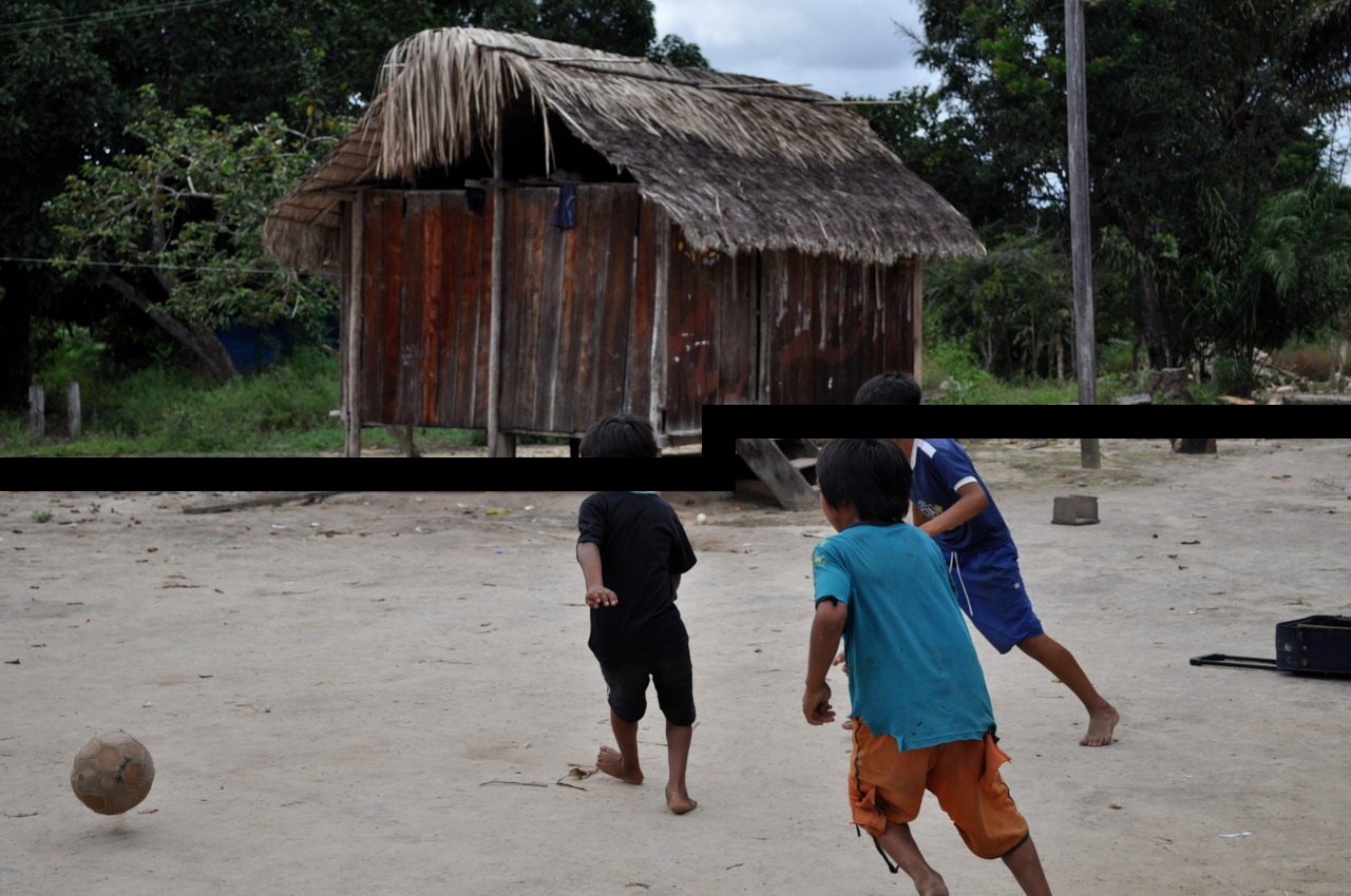 Two children playing football on sandy ground in front of a wooden hut with a thatched roof.