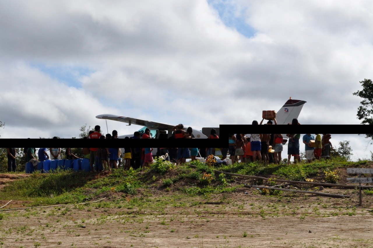 People gathered around a small plane on a grass airstrip, carrying supplies, under a cloudy sky.