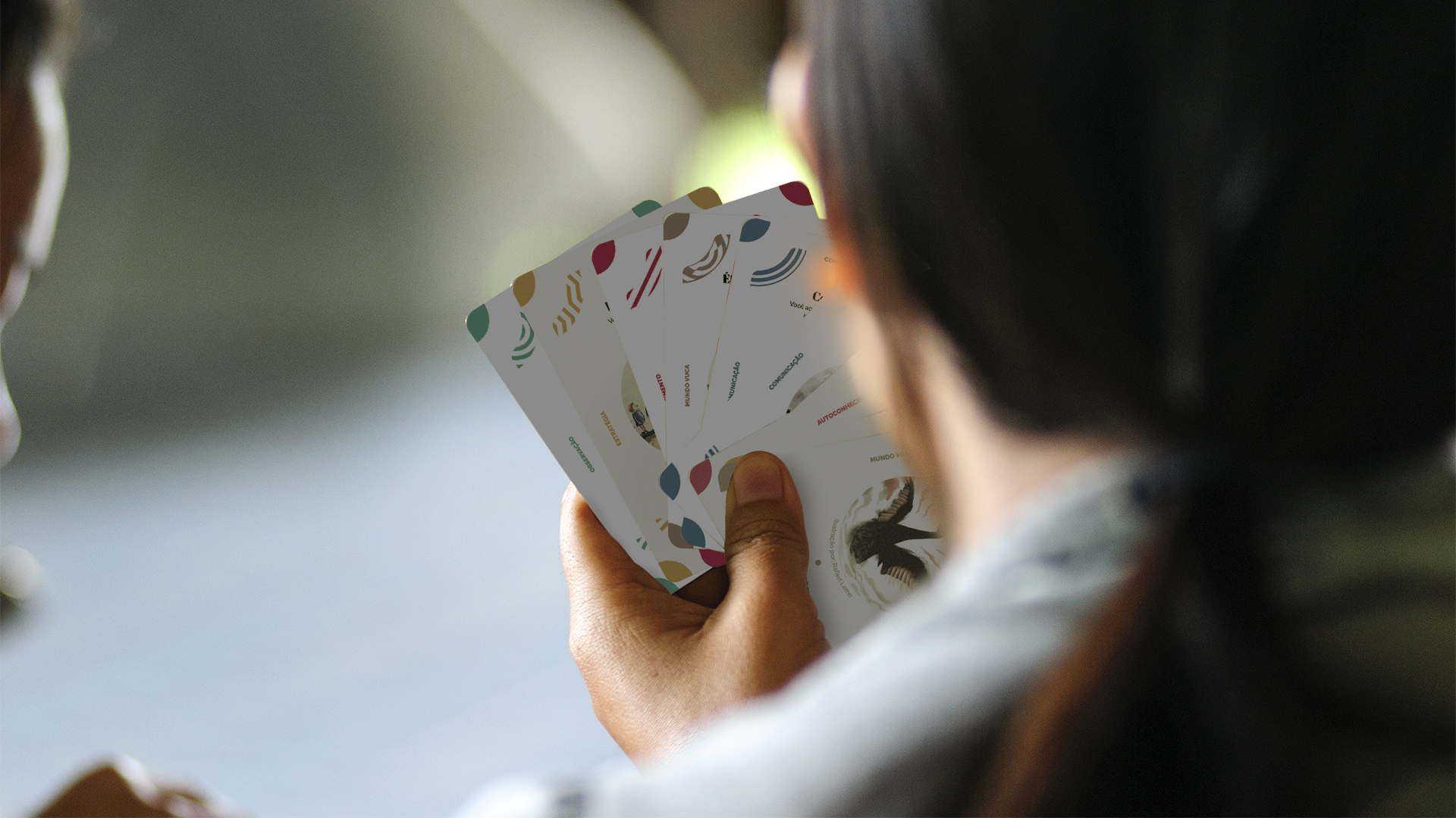 A woman seen from behind holds a fan of illustrated workshop cards, with a soft blurred background.